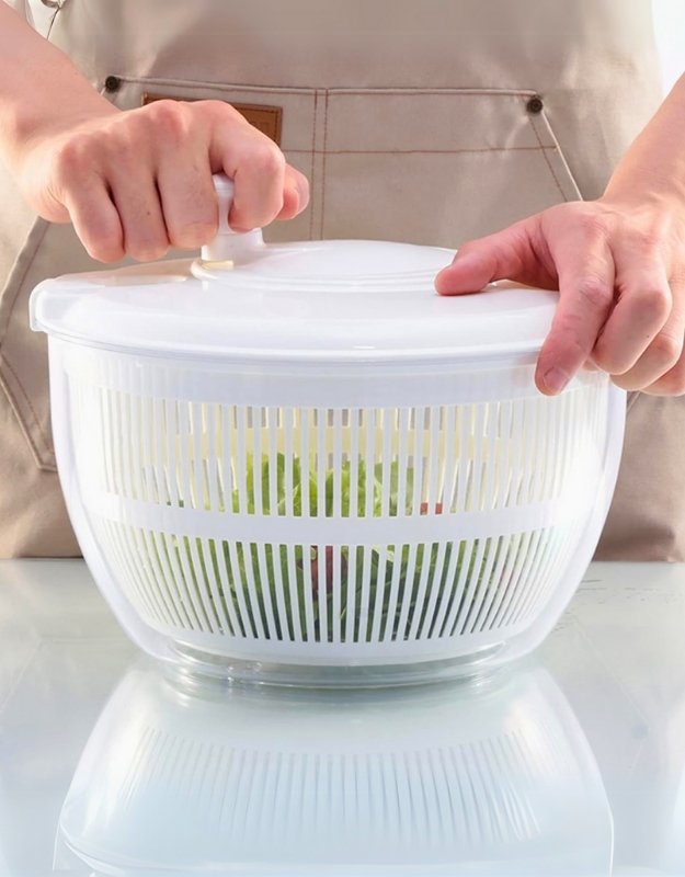 A person is using a white salad spinner to dry vegetables. The spinner is made of plastic and appears to be about 5 liters in capacity.